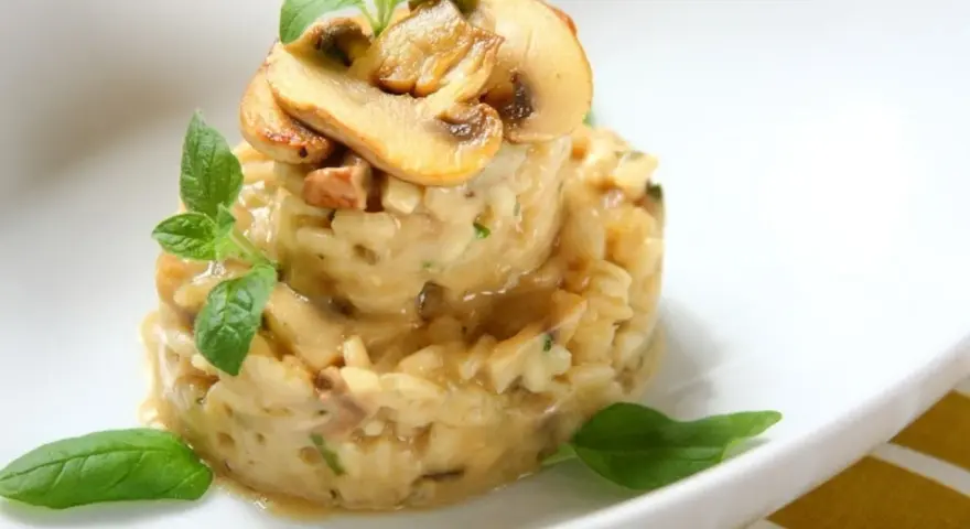 Kremowe risotto z grzybami, udekorowane listkami bazylii i oregano, prezentuje się apetycznie na białym talerzu.