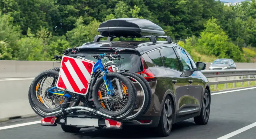 Auto in autostrada con due biciclette sul retro e box sul tetto. Il carico sporgente è ben visibile, pronto per un'avventura.