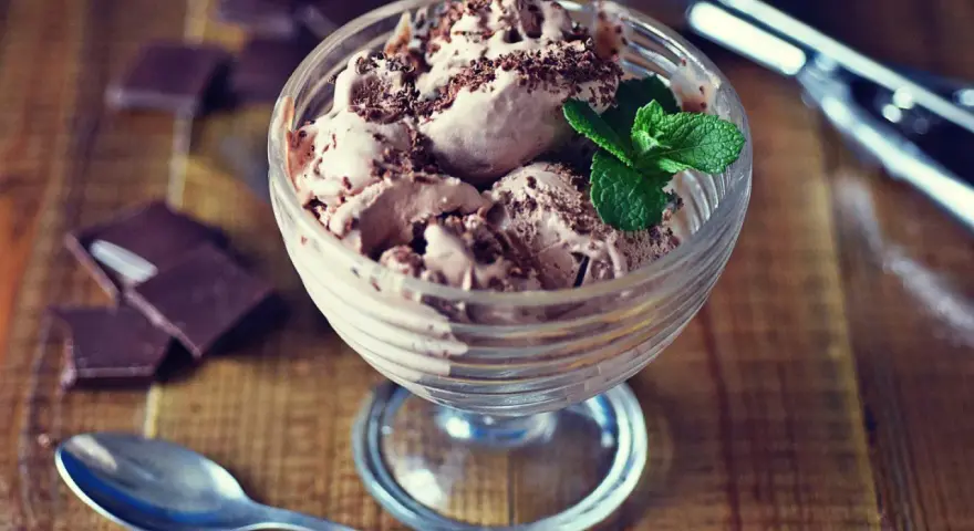 Pyszne lody nuii w pucharku, posypane czekoladą i udekorowane miętą. Obok leżą kawałki czekolady i łyżeczka.