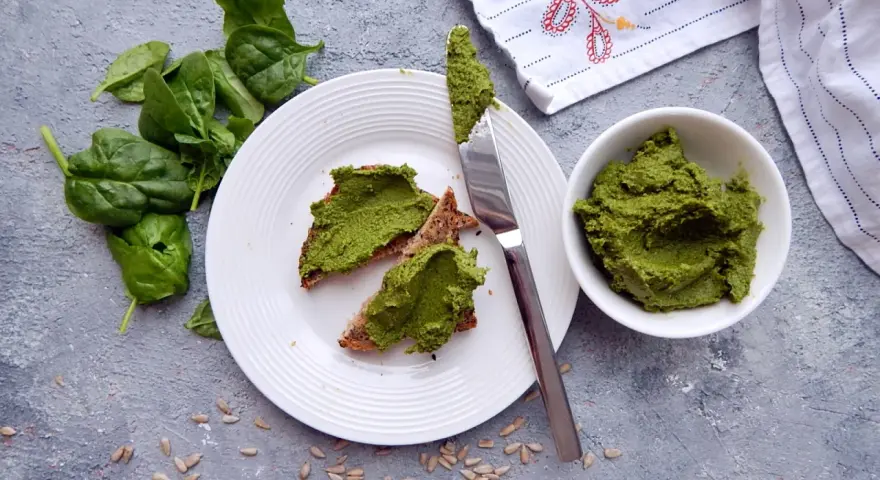 Pesto ze szpinaku do słoików na chlebie z pestkami słonecznika. Świeży szpinak obok talerza.