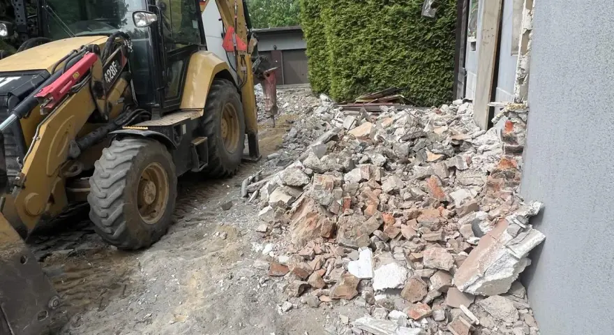 Koparka pracuje przy rozbiórce domu. Gruz i cegły leżą na ziemi, szacowany koszt rozbiórki domu jest wysoki.