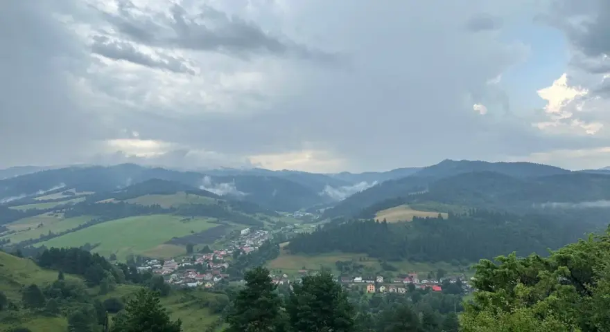 Malownicza panorama z widokiem na wieś w dolinie, otoczoną zielonymi wzgórzami i lasami. Nadciągają chmury, tworząc dramatyczne niebo nad lesnickie sedlo.