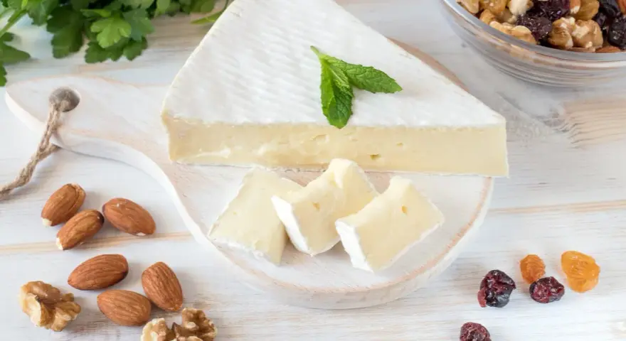 Queso brie en cuña y trozos, decorado con menta. Alrededor hay almendras, nueces y frutos secos. Ideal para quienes buscan información sobre queso brie calorias.