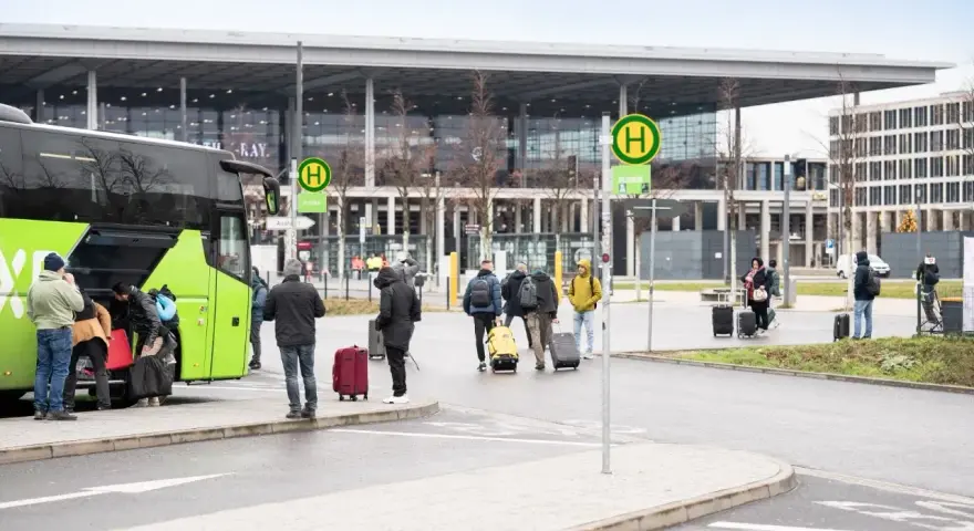 Ludzie z bagażami wsiadają do zielonego autokaru przed nowoczesnym budynkiem, przypominającym lotniska w Berlinie.