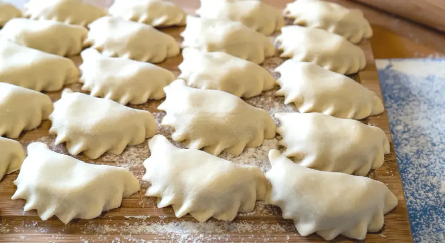 Surowe pierogi ułożone rzędami na drewnianej desce, posypanej mąką.