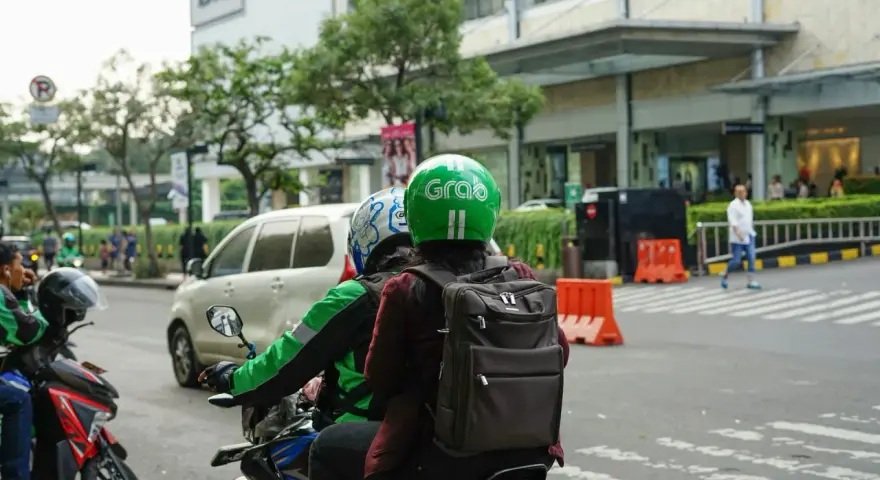 Zwei Personen auf einem Roller, der Fahrer trägt einen grünen Helm mit dem "Grab" Logo. Sie sind unterwegs in Thailand, um mit der Grab App Kunden zu transportieren.