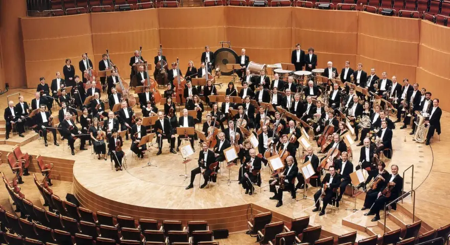Ein großes Orchester mit vielen Musikern, die klassische Instrumente wie Violinen, Celli und Blasinstrumente spielen, posiert auf einer Bühne.
