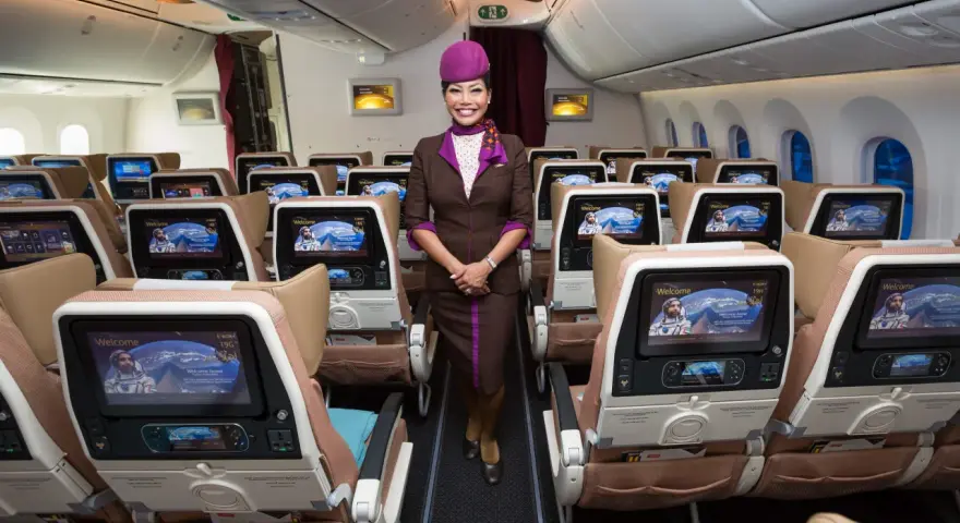 Uśmiechnięta stewardessa Etihad Airways w brązowym mundurku stoi w rzędzie foteli. Ekraniki pokazują astronautę. Dobre etihad airways opinie.