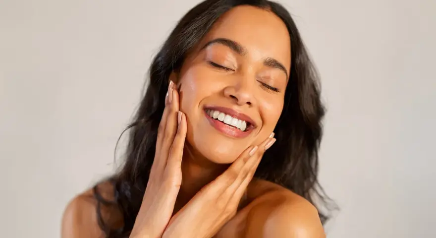 Une femme au sourire radieux caresse son visage, révélant une peau glowy éclatante de santé.
