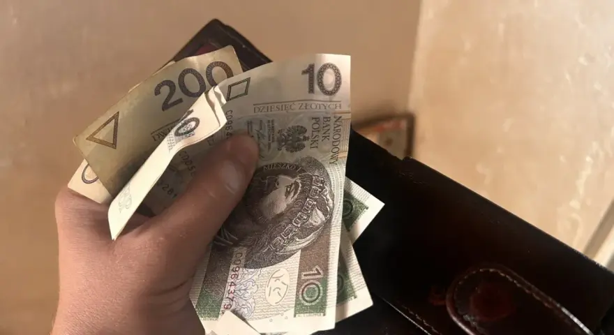 Dłoń trzyma polskie banknoty 10 zł i 200 zł obok portfela.