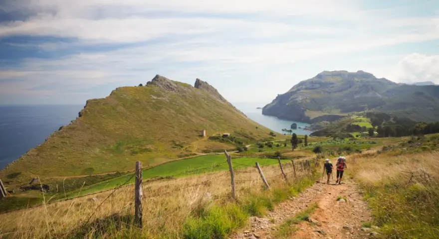 Two hikers with backpacks walk on a dirt path through a grassy landscape with mountains and the sea in the background.