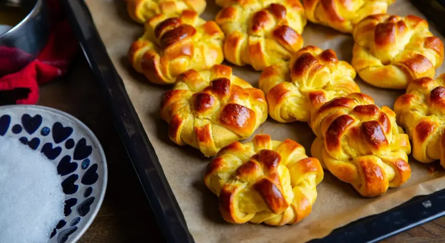 Nybakade lussebullar med kesella, gyllene och redo att avnjutas. En skål med socker och en röd duk anas i bakgrunden.