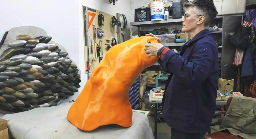 Artist applies finishing touches to a large, orange, 3D printed object, part of a project creating large objects.