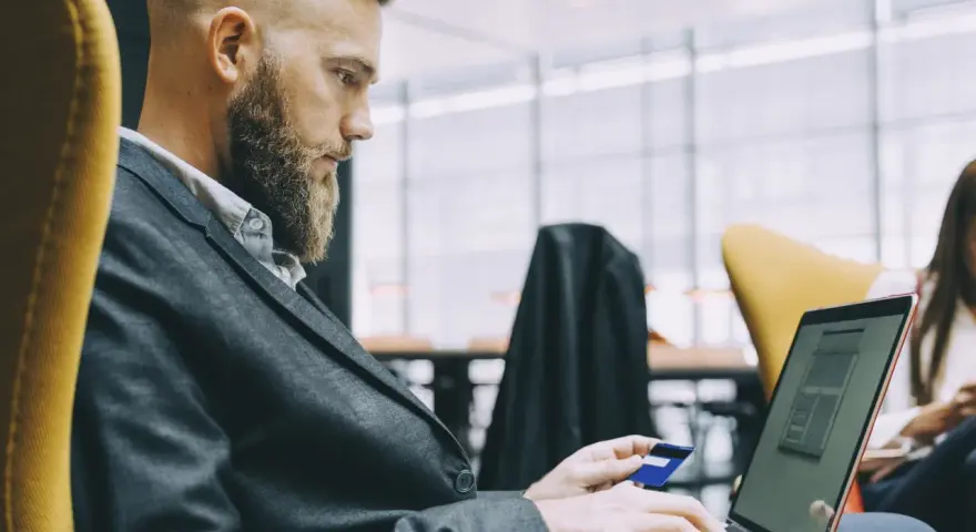 Hombre con barba y traje usa tarjeta de crédito en portátil. Busca cuánto se paga la hora extra en España.