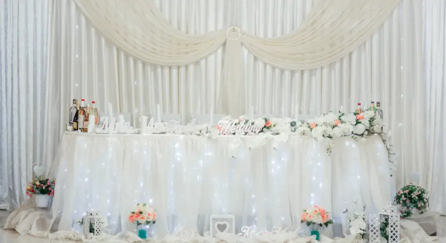 Elegancka ścianka za parą młodą inspiracje: białe zasłony, kwiaty, napisy "Wedding", "Love", "Mrs & Mr".