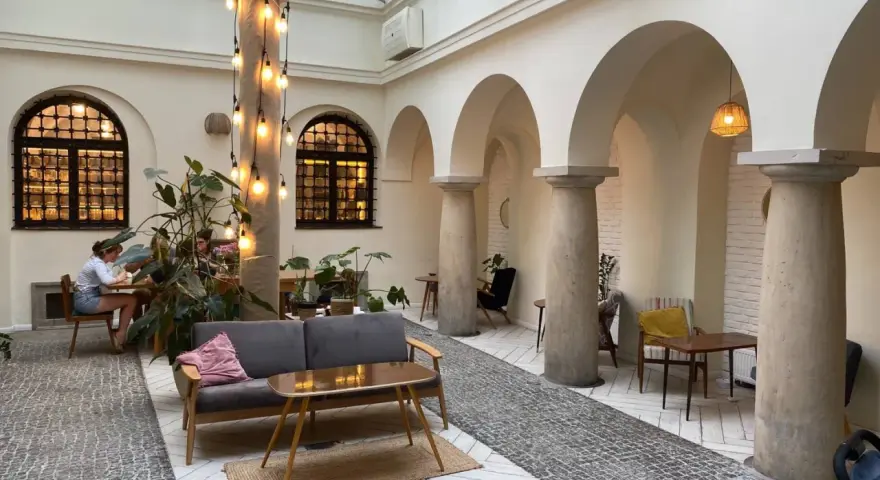 Przytulna kawiarnia patio z łukami i kolumnami. Goście siedzą przy stolikach, a obok stoi wygodna sofa. Widać rośliny i dekoracyjne lampki. Menu czeka na odkrycie.