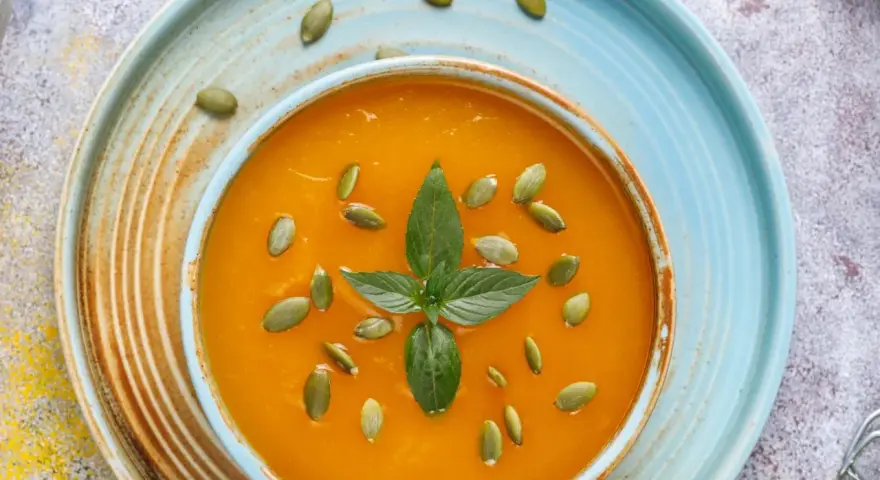 Zupa krem z dyni przepis Ewy Wachowicz. Aromatyczna, pomarańczowa zupa z pestkami dyni i listkiem mięty na niebieskim talerzu.