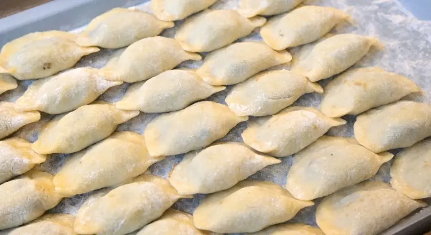 Ułożone na tacy surowe pierogi, posypane mąką.