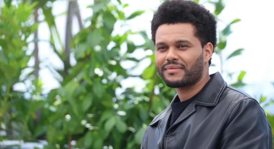 The Weeknd trägt eine schwarze Lederjacke vor einem grünen, unscharfen Hintergrund.