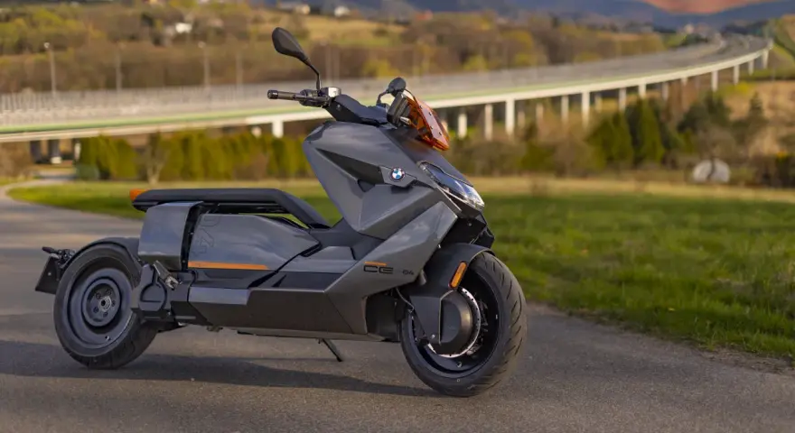 Szary skuter elektryczny BMW CE 04 na tle malowniczego krajobrazu z mostem i wzgórzami.