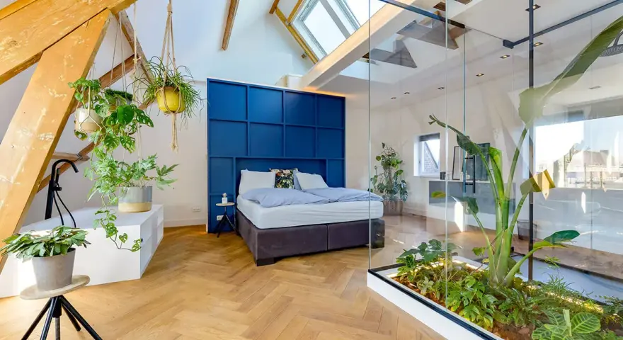 Mansarde da sogno con letto blu, piante sospese e un giardino interno in vetro. Travi a vista e lucernari creano un'atmosfera unica.