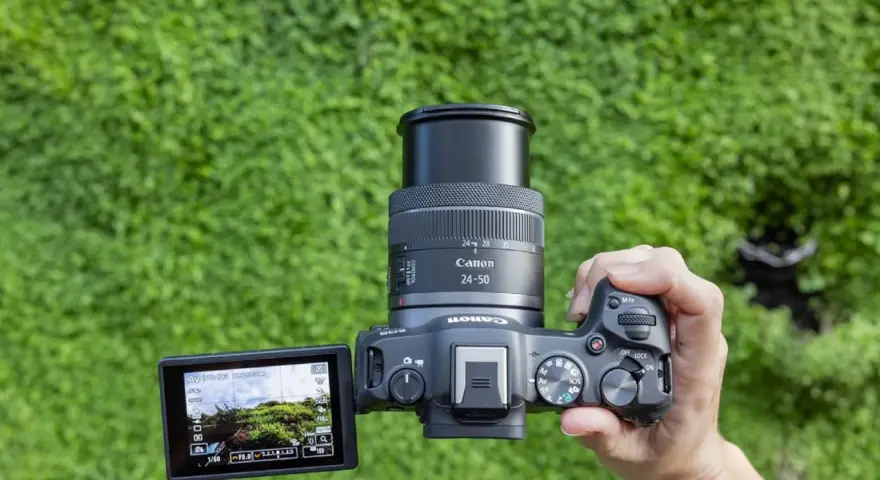 Main d'une personne tenant un appareil photo Canon R8 avec un objectif 24-50mm, prêt pour un test. L'écran affiche un paysage verdoyant.