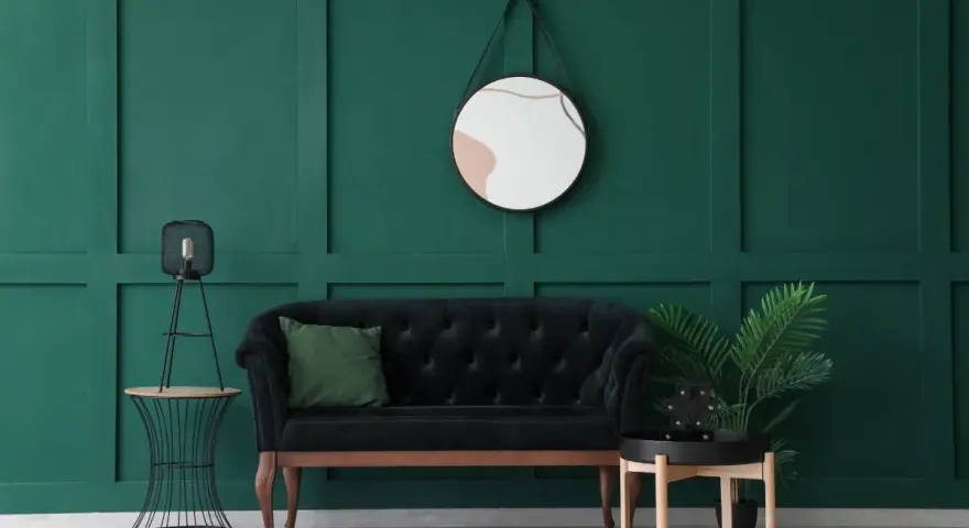 Un elegante divano nero con cuscino verde, un tavolino con lampada e una pianta, e uno specchio dietro il divano su una parete verde scuro.