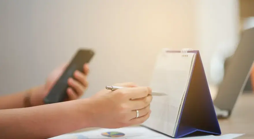 Ręce planują zadania na kalendarzu, trzymając telefon. Zapewnia to ciągłość pracy i organizację dnia.