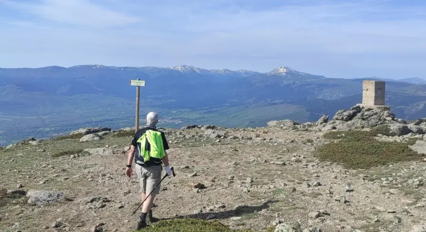 Senderista camina hacia el **puerto de Navafría**, con un mojón y montañas nevadas al fondo.