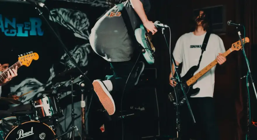 Ein Gitarrist springt auf der Bühne, die Energie des Punk der 70er Jahre ist spürbar. Ein Bassist spielt im Hintergrund.