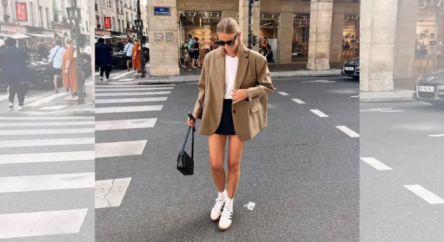Mujer con look con blazer beige, falda negra y zapatillas blancas cruza un paso de peatones en París.