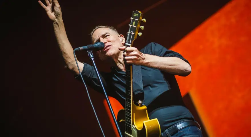Bryan Adams na koncercie, w czarnej koszuli, z gitarą i mikrofonem, wznosi rękę w geście pozdrowienia.