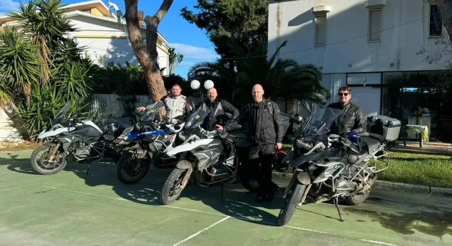 Czterech motocyklistów w pełnym rynsztunku gotowych na **prom na Sycylię**. Motocykle BMW GS zaparkowane na zielonym placu, w tle budynek i palmy.