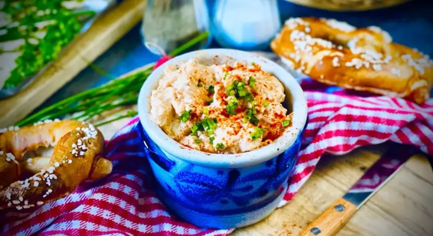 Herzhaftes Obazda-Rezept in einem blauen Steingut-Töpfchen, garniert mit Schnittlauch und Paprikapulver, daneben Brezeln und ein Messer.