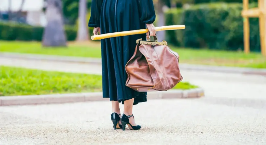 Elegancka kobieta w czarnej sukni i czarnych szpilkach, niosąca brązową torbę i żółty kij. Uosobienie francuskiej mody.