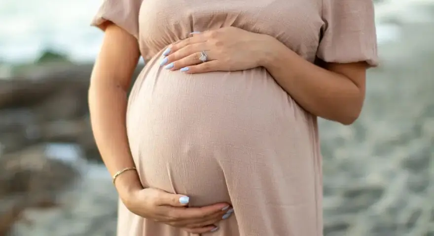 Brzuszek ciężarnej kobiety w beżowej sukience, dłonie delikatnie go obejmują. Warto pamiętać o bezpieczeństwie i unikać stosowania **retinolu w ciąży**.