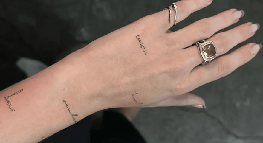 Delikatny tatuaż "famiglia" na prawej dłoni, obok pierścionków. Na lewej ręce widnieje napis "Amour" i "sandala".