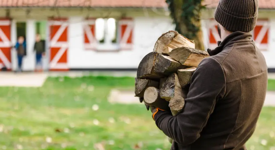 Chcesz wiedzieć, jak zostać leśniczym? Ten mężczyzna z drewnem na podpałkę, w tle dom z czerwonymi okiennicami i dzieci, pokazuje, że praca w lesie to też praktyczne zadania.