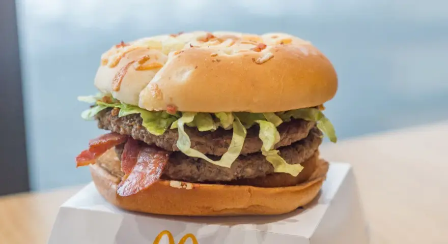 Duży burger z dwoma kotletami, bekonem i sałatą na białym opakowaniu.