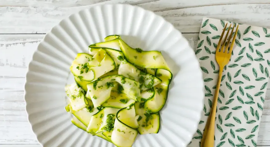 Delicato carpaccio di zucchine condito con erbe fresche, servito su un piatto bianco con forchetta dorata.