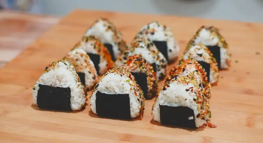 Rząd trójkątnych onigiri z kurczakiem, posypanych sezamem i przyprawami, ułożonych na drewnianej desce.