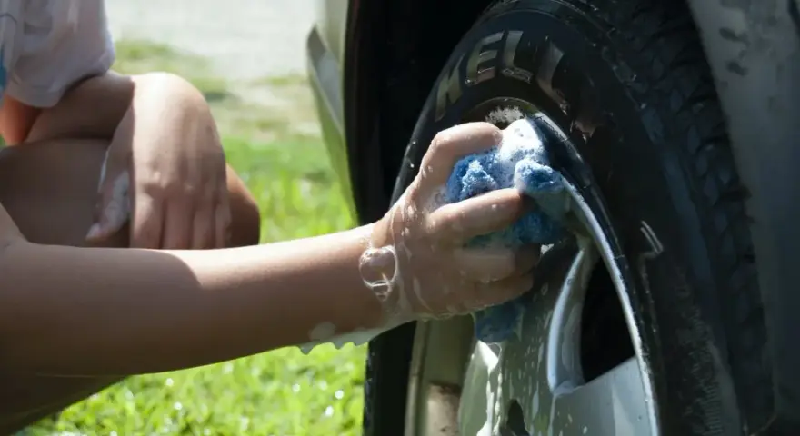 A person's arm and hand, covered in suds, scrub a car tire with a blue sponge. It seems they are testing if you can use dish soap to wash your car.