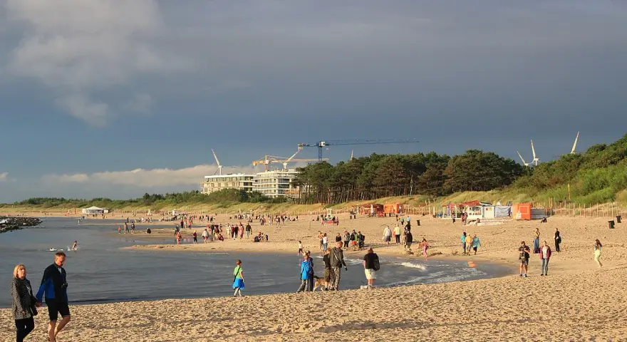 Nadmorskie miasto nad Wieprzą tętni życiem. Ludzie spacerują po plaży, bawią się w wodzie, a w tle widać budowę i wiatraki.
