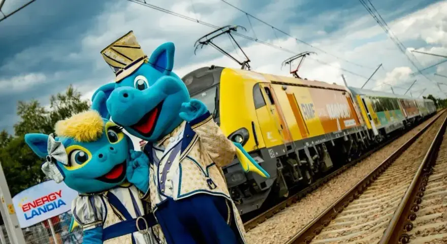 Dwa niebieskie smoki w strojach stoją obok żółtego pociągu na torach kolejowych.