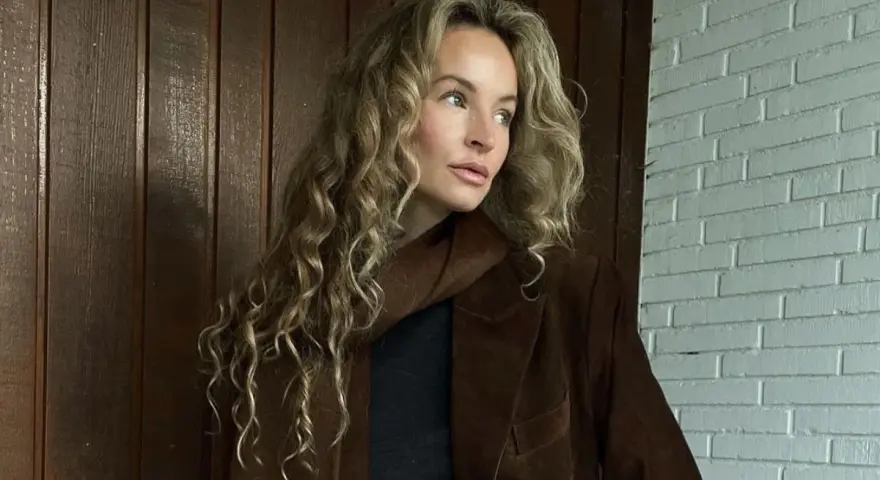 Kobieta z kręconymi, beżowymi włosami, którym pasuje ten odcień. Elegancka stylizacja z brązową marynarką.