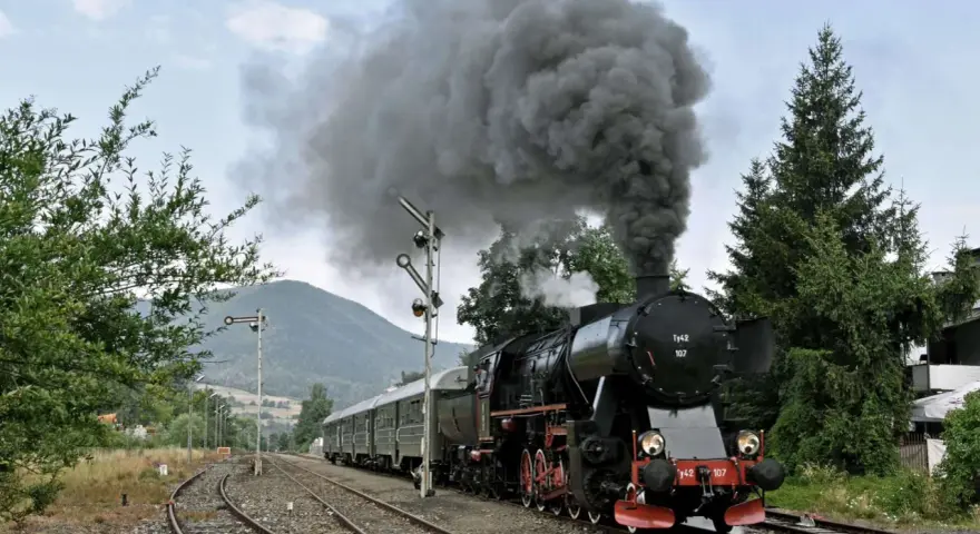 Czarny, parowy pociąg retro z wagonami, wydobywający kłęby dymu, jedzie po torach wśród zieleni i gór.