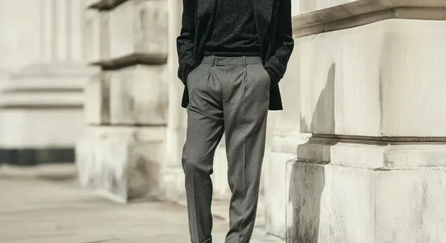 Uomo con outfit elegante: giacca scura, maglione nero e pantaloni chino grigi. Scarpe stringate lucide completano il look.