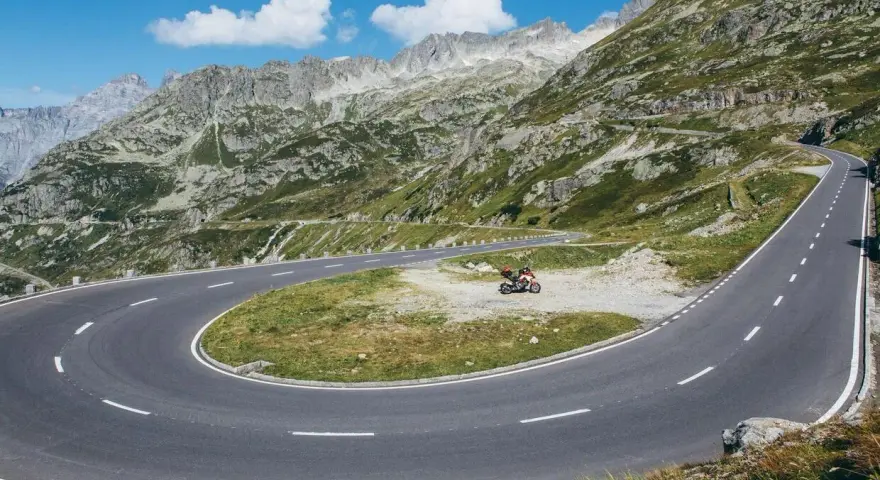 Una moto attende su un tornante del Grimsel Pass, tra montagne verdi e cielo azzurro.