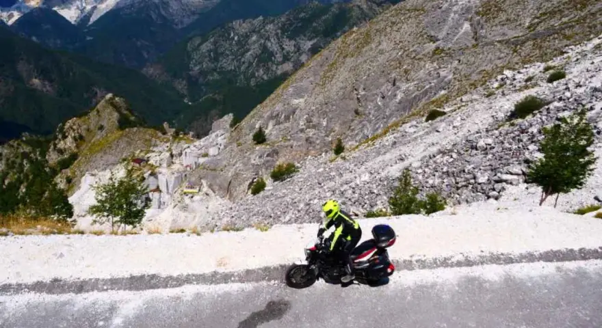 Motociclista in abbigliamento giallo fluo affronta il passo del vestito, tra cave di marmo e montagne imponenti.
