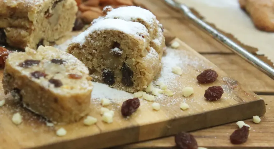 Stollen formen ohne backform: saftige Stollenstücke mit Rosinen und Puderzucker auf Holzbrett, umgeben von Nüssen und Trockenfrüchten.